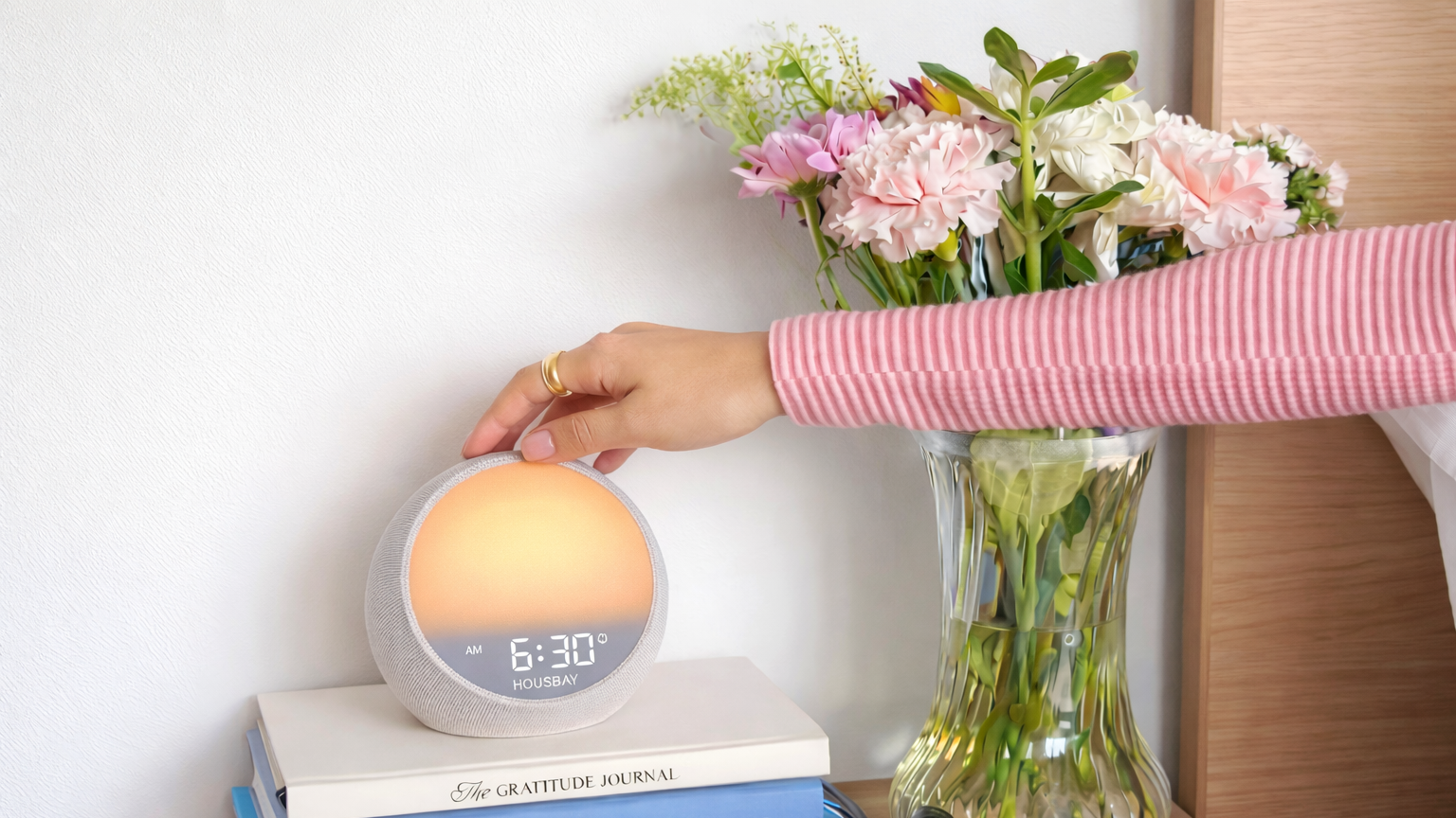 Person adjusting a sunrise alarm clock on a stack of books with flowers in the background