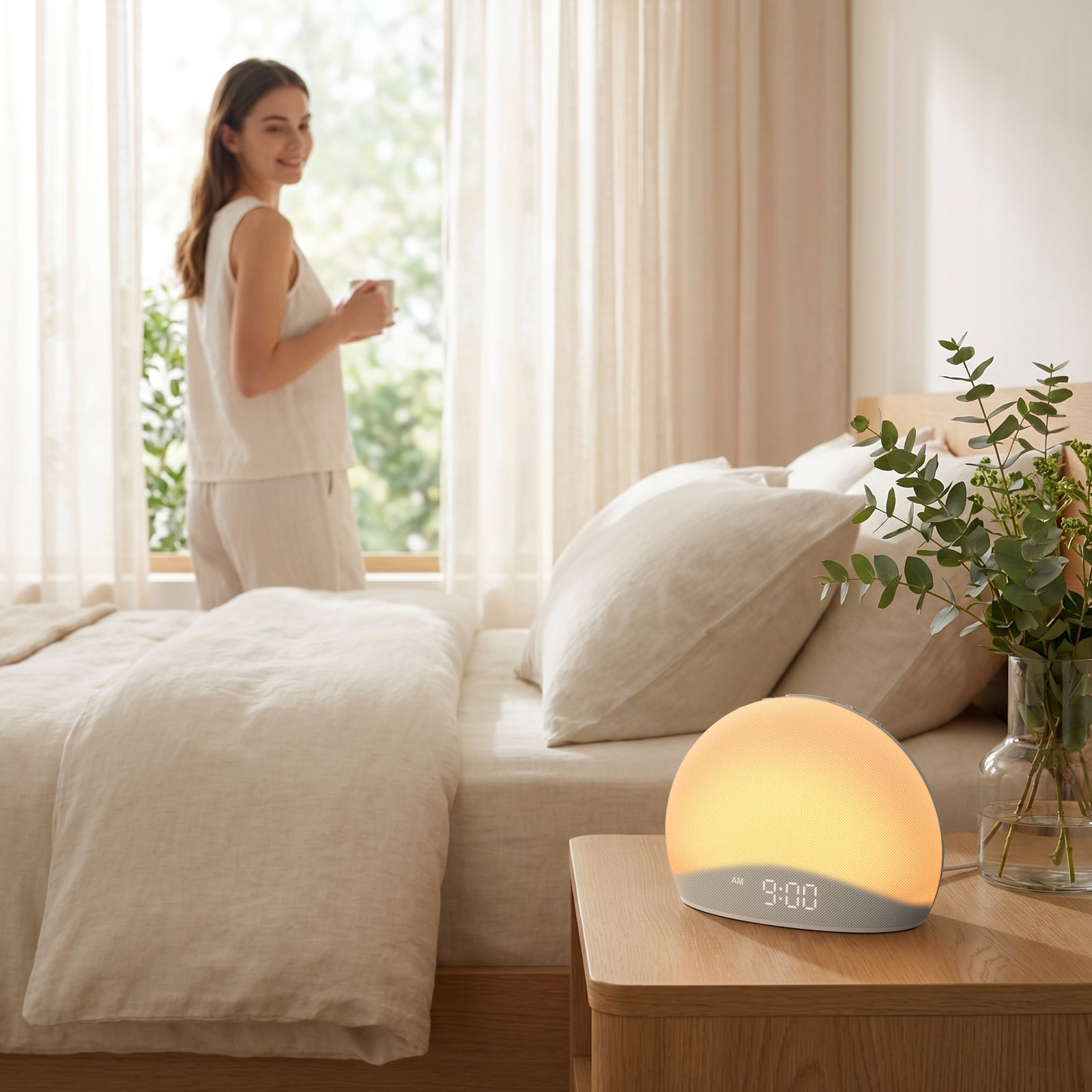 A woman standing by the window holding a mug in a bright, cozy bedroom, with the Housbay Dawn wake-up light glowing gently on the nightstand at 9:00 AM.