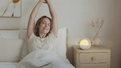Woman waking up gently in a sun-lit bedroom with the Rhythmana MINI sunrise alarm clock glowing softly on the nightstand.