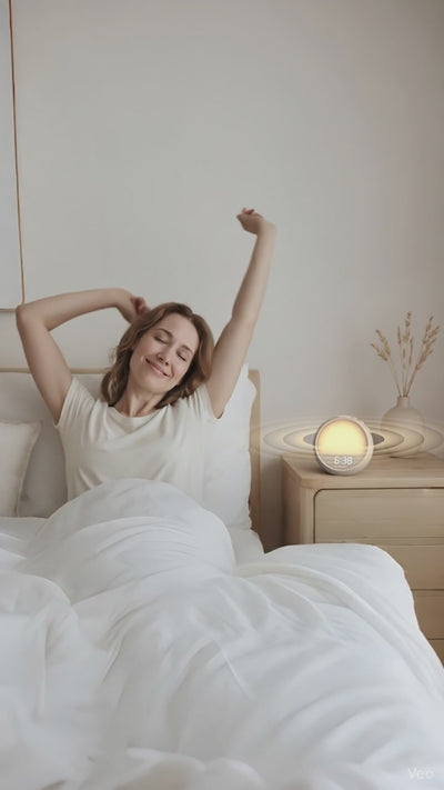 Woman waking up gently in a sun-lit bedroom with the Rhythmana MINI sunrise alarm clock glowing softly on the nightstand.